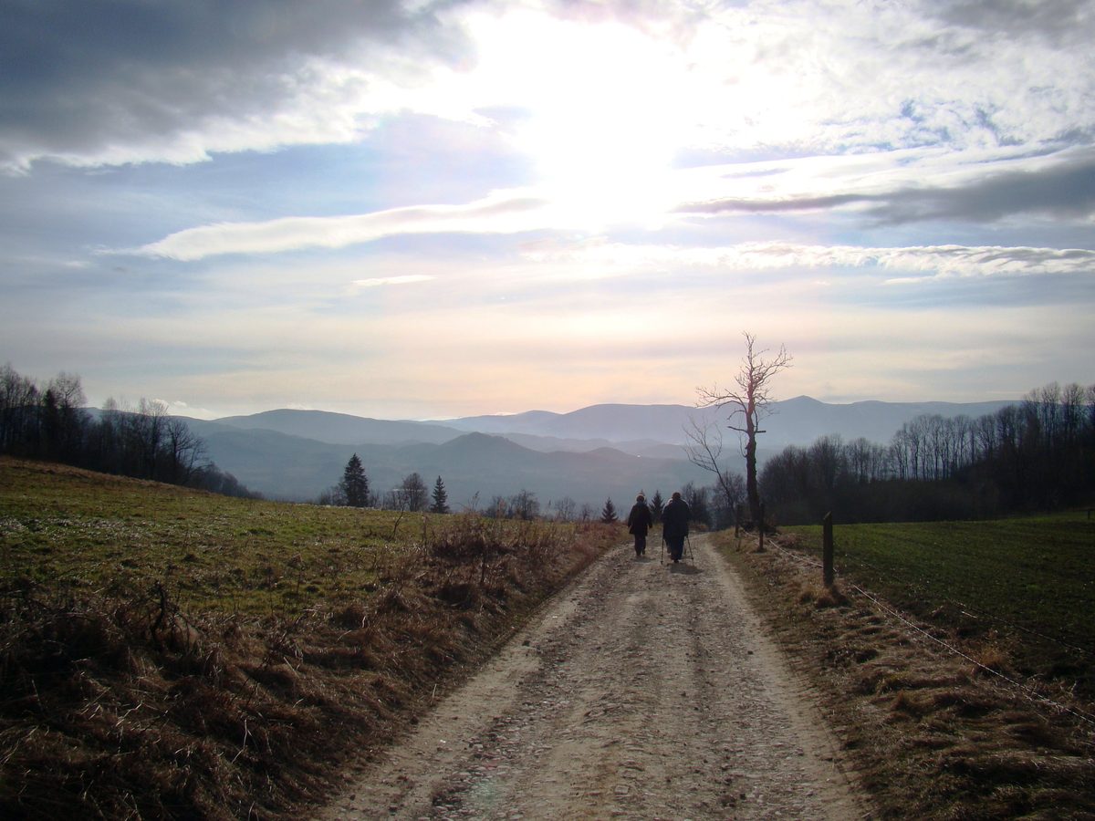 Piaszczysto-kamienista droga gruntowa biegnąca grzbietem, dwie sylwetki idących z kijkami trekkingowymi, w tle warstwy pasm górskich pod jasnym, częściowo zachmurzonym niebem.