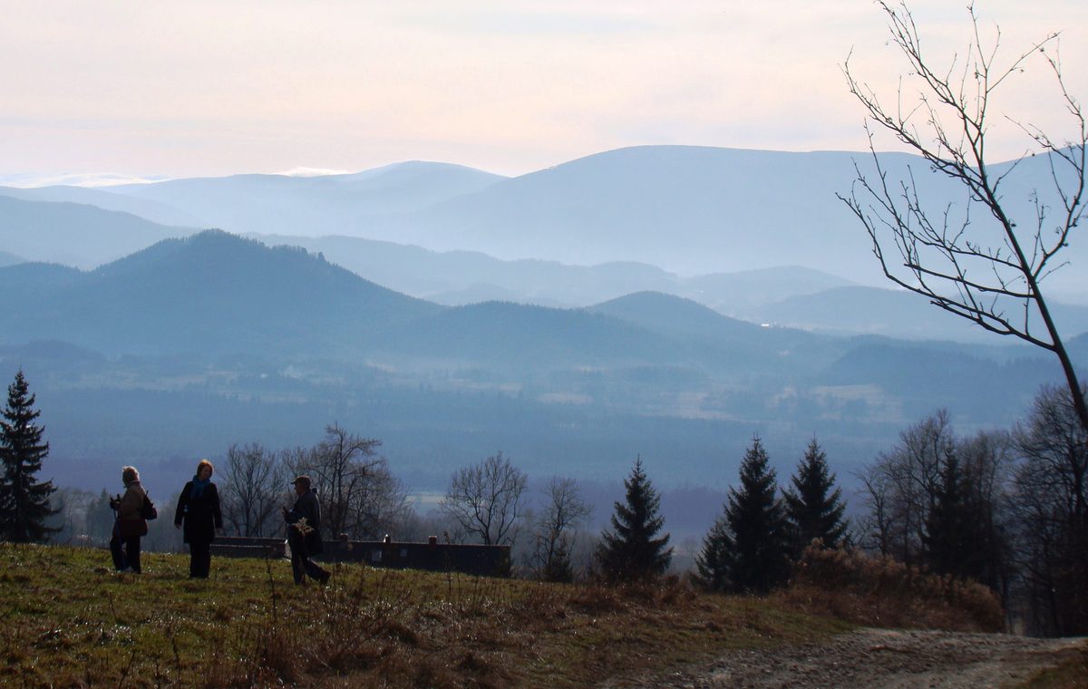 Szeroka panorama warstwowych, zamglonych pasm górskich widziana znad otwartego zbocza, na pierwszym planie trzy osoby idące łąką i uschnięte drzewo.