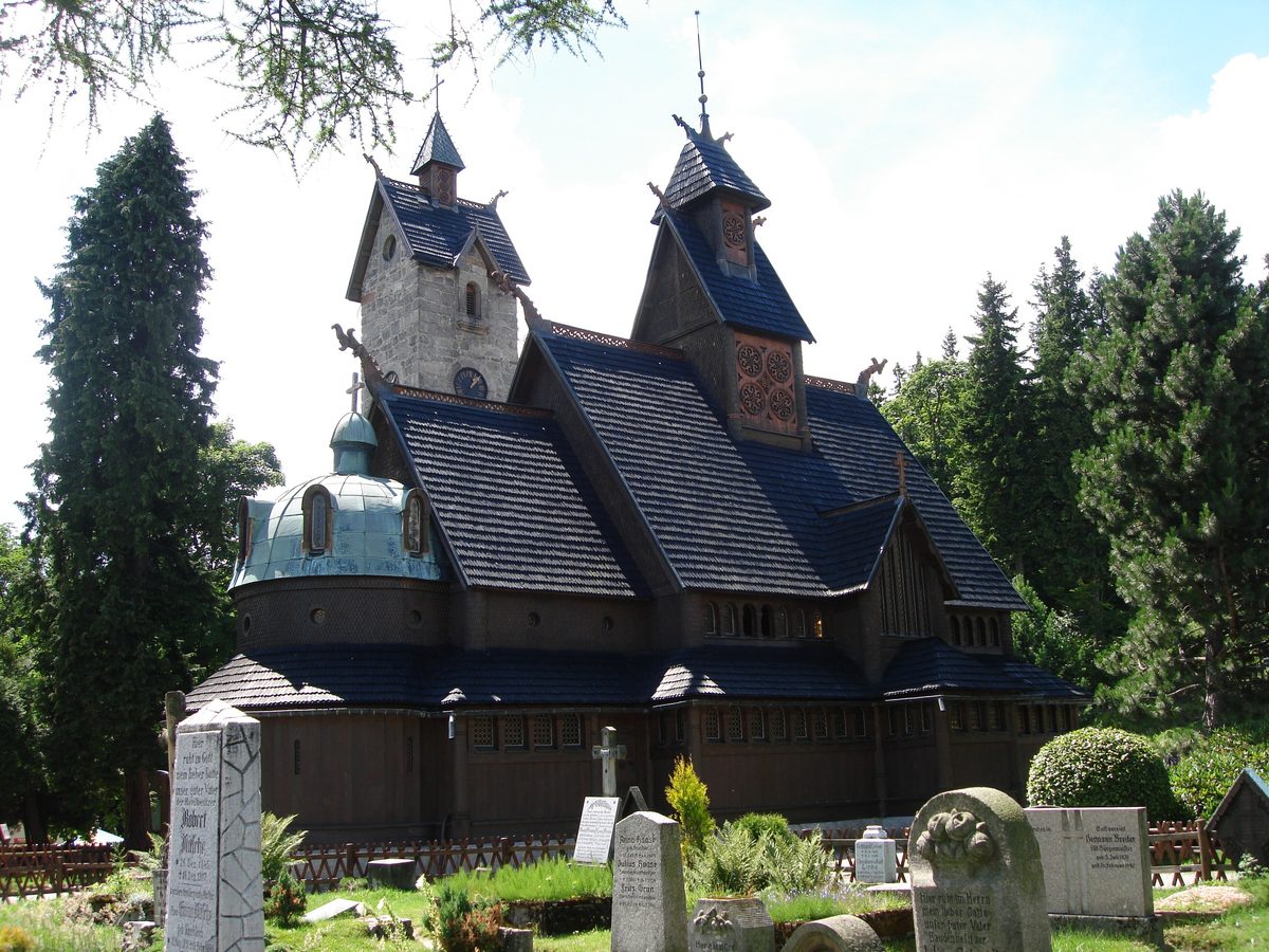 Drewniany kościół z zieloną kopułą i kamienną wieżą w słońcu, dachy w chłodnym odcieniu, wokół świerki i rzędy starych nagrobków.