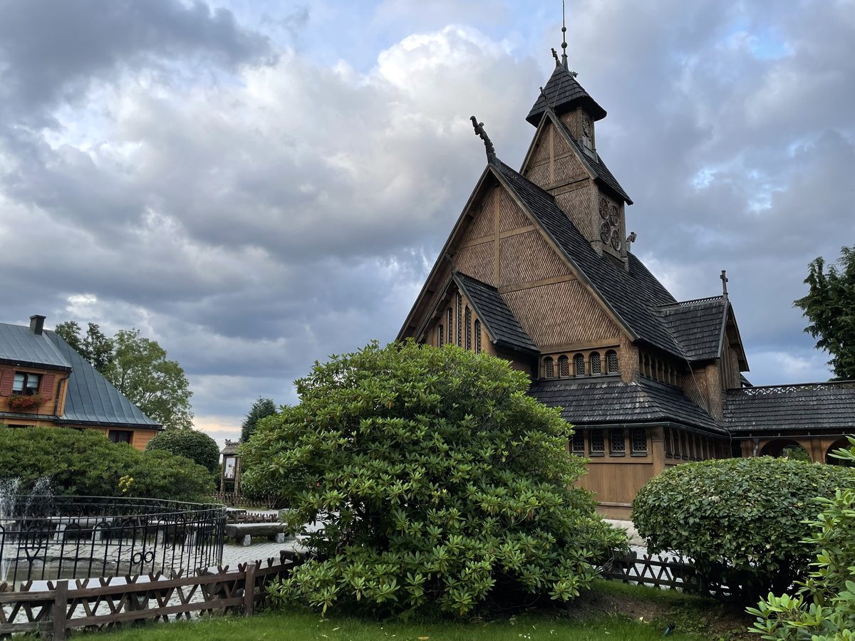 Drewniany kościół z wysokim gontowym dachem i sygnaturką na tle burzowych chmur, przed nim strzyżone krzewy i drewniane ogrodzenie fontanny.
