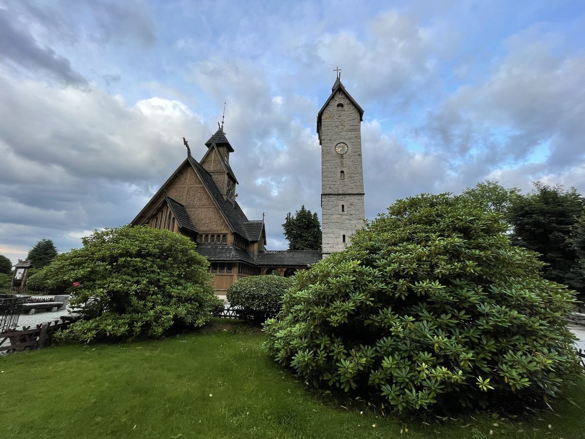 Drewniany kościół i kamienna wieża zegarowa nad żywopłotem z rododendronów, w tle grzbiet gór, niebo w chmurach o zmierzchu.