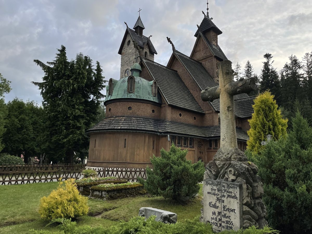 Drewniany kościół z zieloną kopułą i kamienną wieżą, przed nim kamienny krzyż nagrobny i strzyżone krzewy, pochmurne niebo.