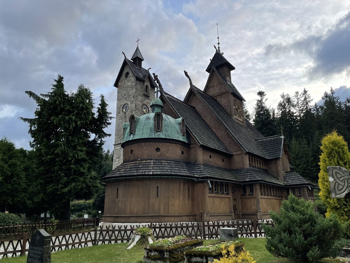 Drewniany kościół z zieloną miedzianą kopułą, kamienną wieżą zegarową i stromymi gontowymi dachami, wokół świerki i drewniane ogrodzenie.
