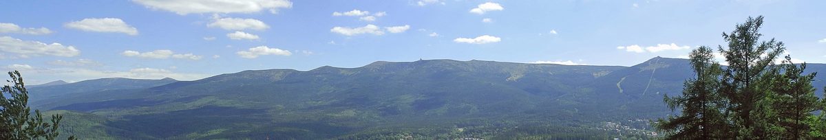 Panorama grzbietu Karkonoszy z Drogi Sudeckiej nad Szklarską Porębą: w dole zalesione zbocza i zabudowa miasta, na horyzoncie łagodna, długa grań pasma, po prawej stoki narciarskie pod Szrenicą
