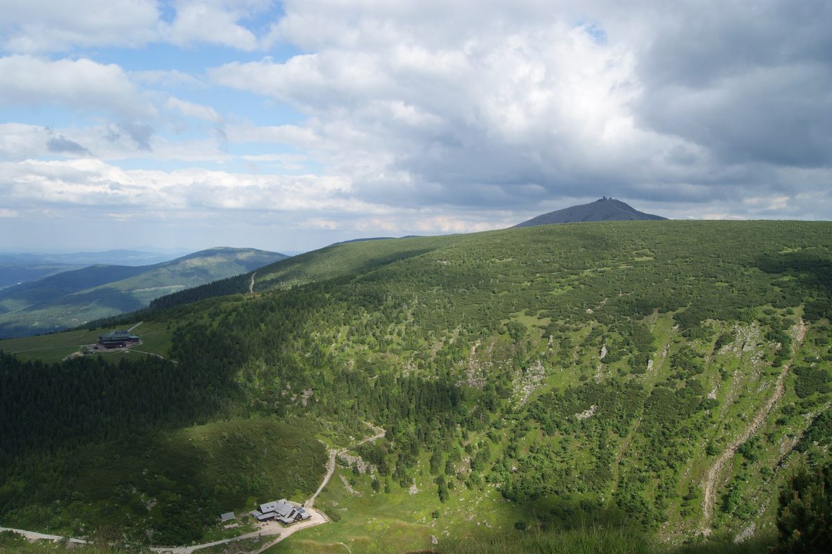 Szeroka panorama zielonego trawiastego grzbietu z kopułą szczytu w tle, w dolinie dwa niewielkie budynki, chmurzące się niebo, lato.