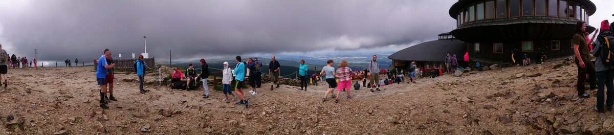 Panorama kamienistego plateau z tłumem turystów w kurtkach, po prawej okrągły budynek z dyskowym dachem. Ciężkie szare chmury.