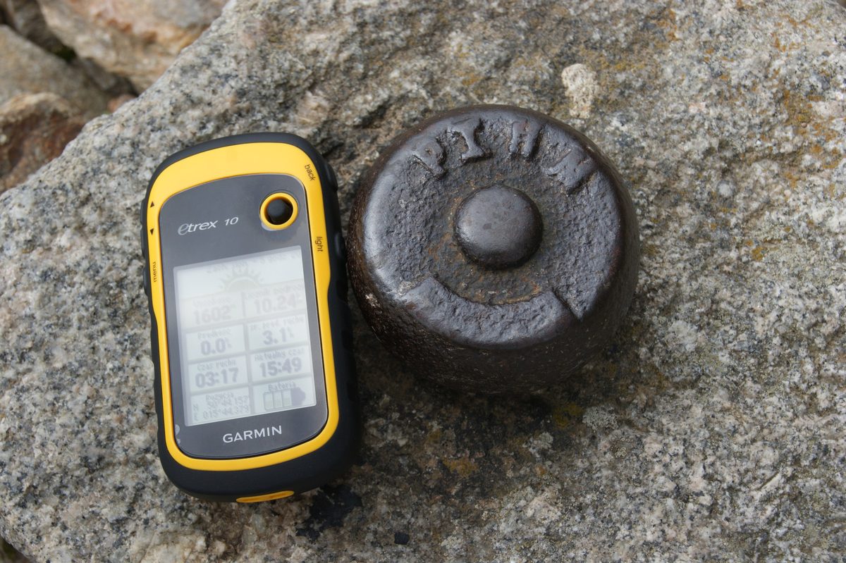 Żółto-czarny GPS Garmin eTrex 10 z odczytem 1602 m leży na granitowej skale obok metalowej głowicy punktu osnowy geodezyjnej. Zbliżenie.