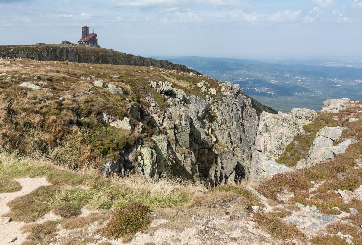 Skalne żebro w pierwszym planie, dalej urwista ściana kotła zakończona budynkiem z czerwonym dachem i wieżą, po prawej równina we mgle.