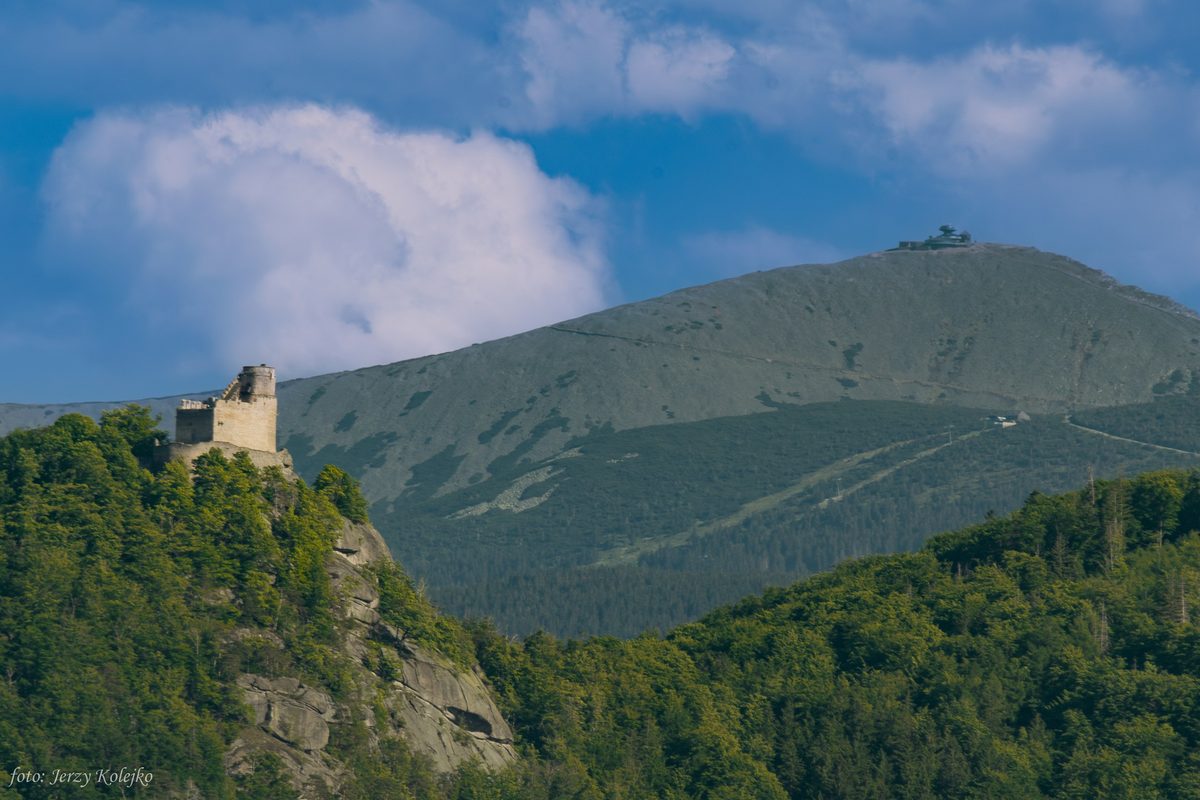 Ruiny zamku z wieżą na skalistym cyplu wśród lasu, w tle rozległy grzbiet górski z obserwatorium na szczycie. Lato.